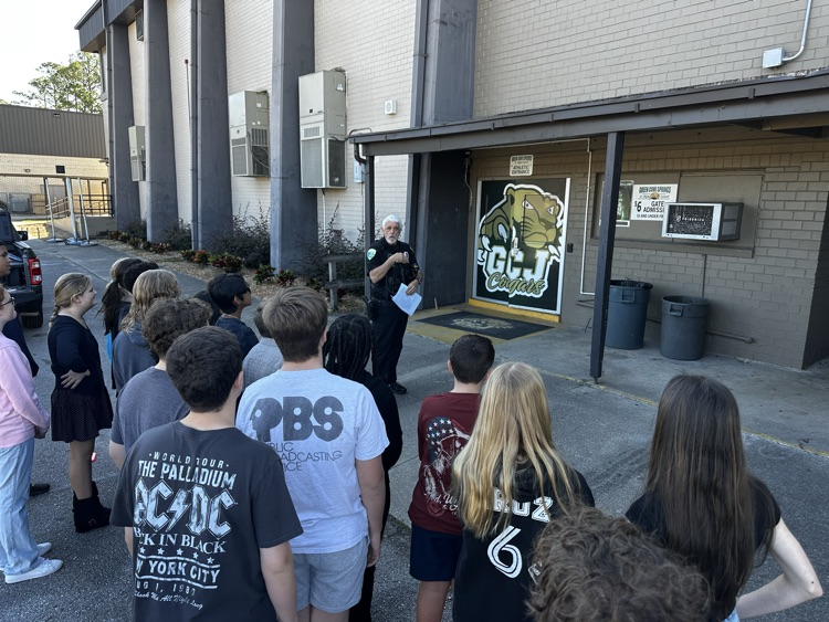 A group of students outside a building. A man stands in front of them. Trees and plants in the background.
