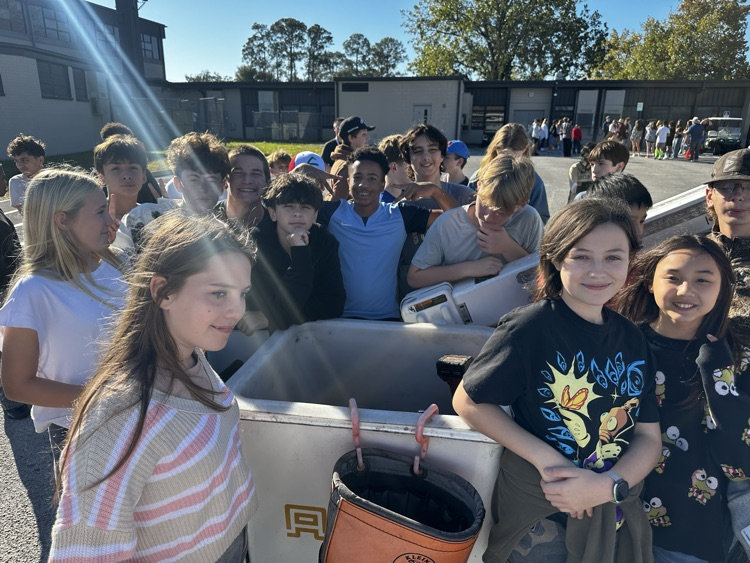 Several young people gather around a large container with a handle, standing outdoors near a building.