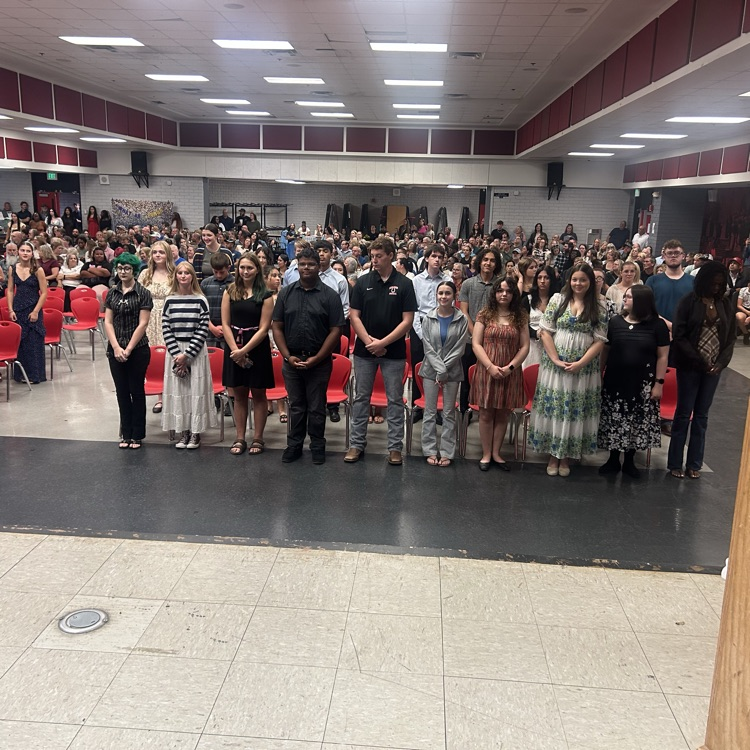 Many people stand in rows on a stage under fluorescent lights. Some people wear dresses, others suits.