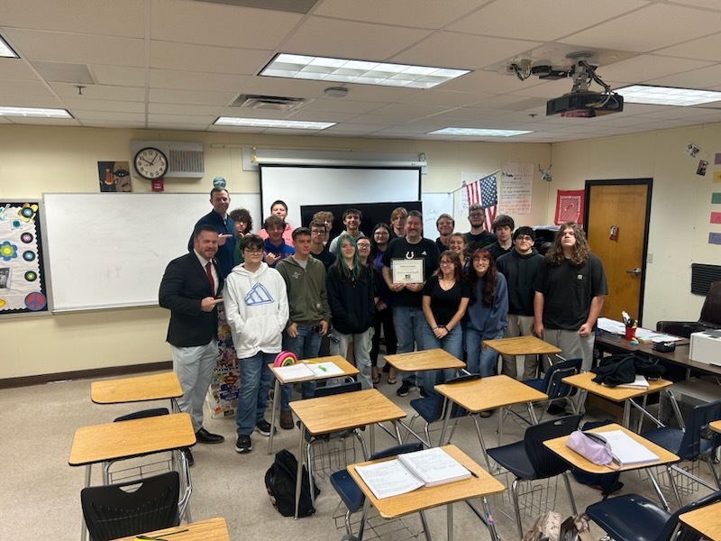A classroom with several students and adults standing. A teacher stands at the front with a group of students.