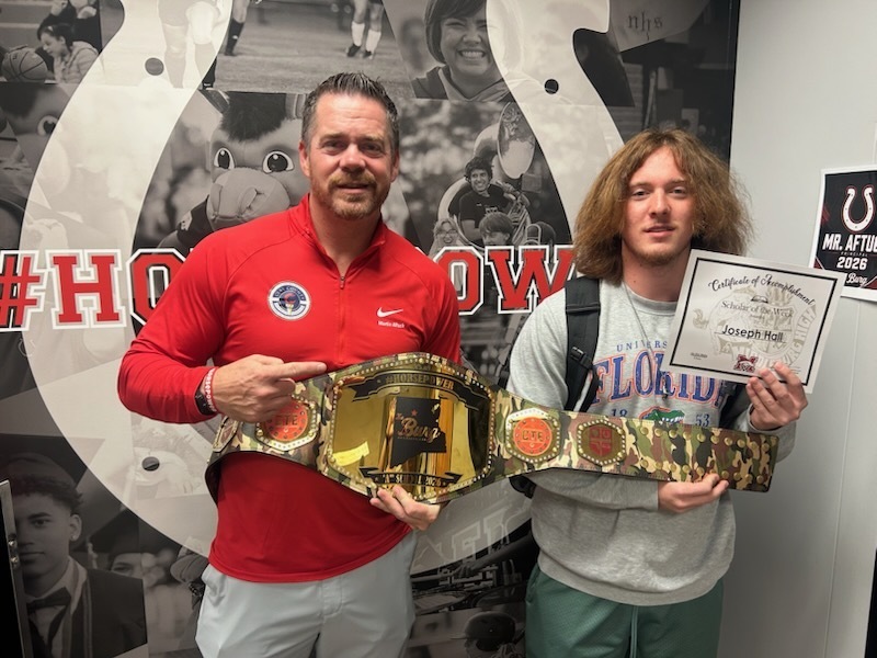 Two people stand in front of a wall with a wrestling belt and a certificate.