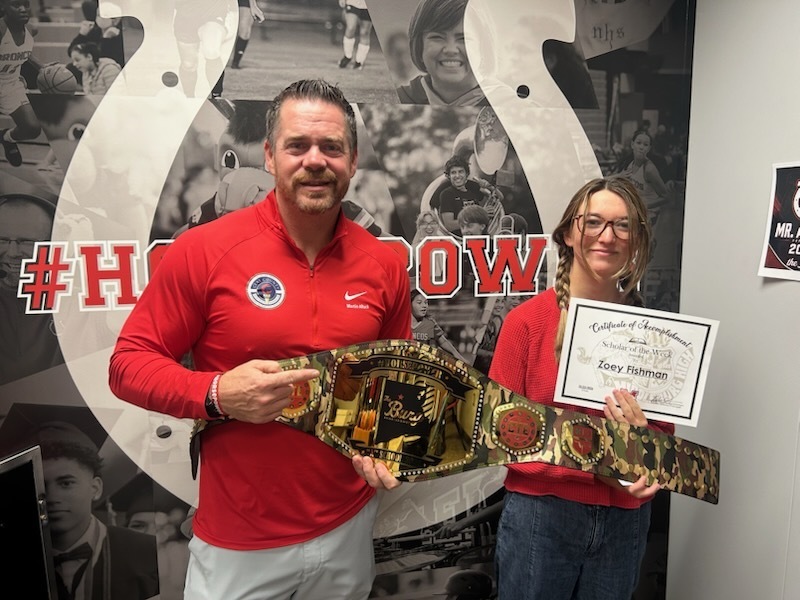 A man and woman in red shirts hold a belt and certificate, standing before a photo of a sports team.