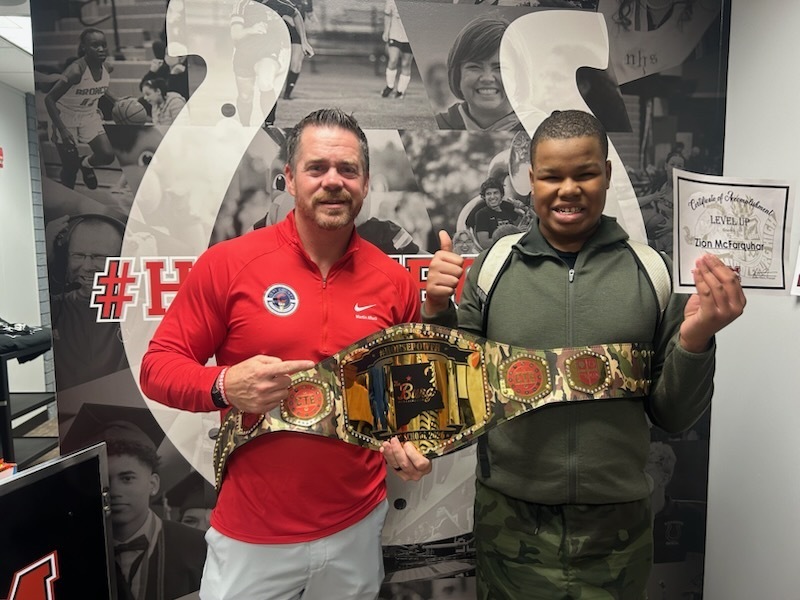 Two men hold a large belt with buckles in front of a wall with sports photos.