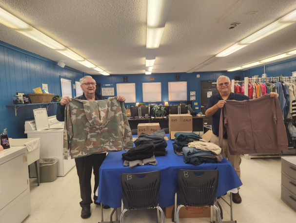 Two men standing at a table hold up articles of clothing. Behind them, a laundry area.