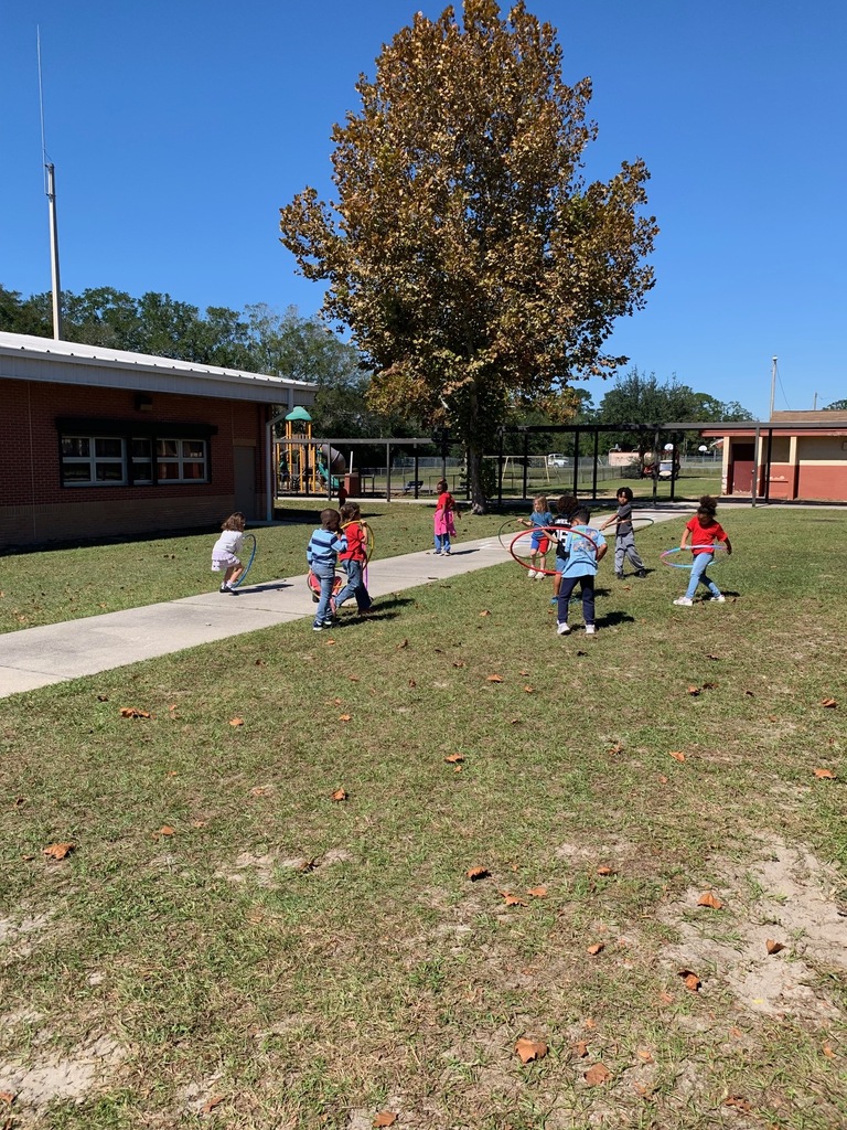 Children walk and play on a grassy lawn near a building with windows. A tree with sparse leaves is in the background.