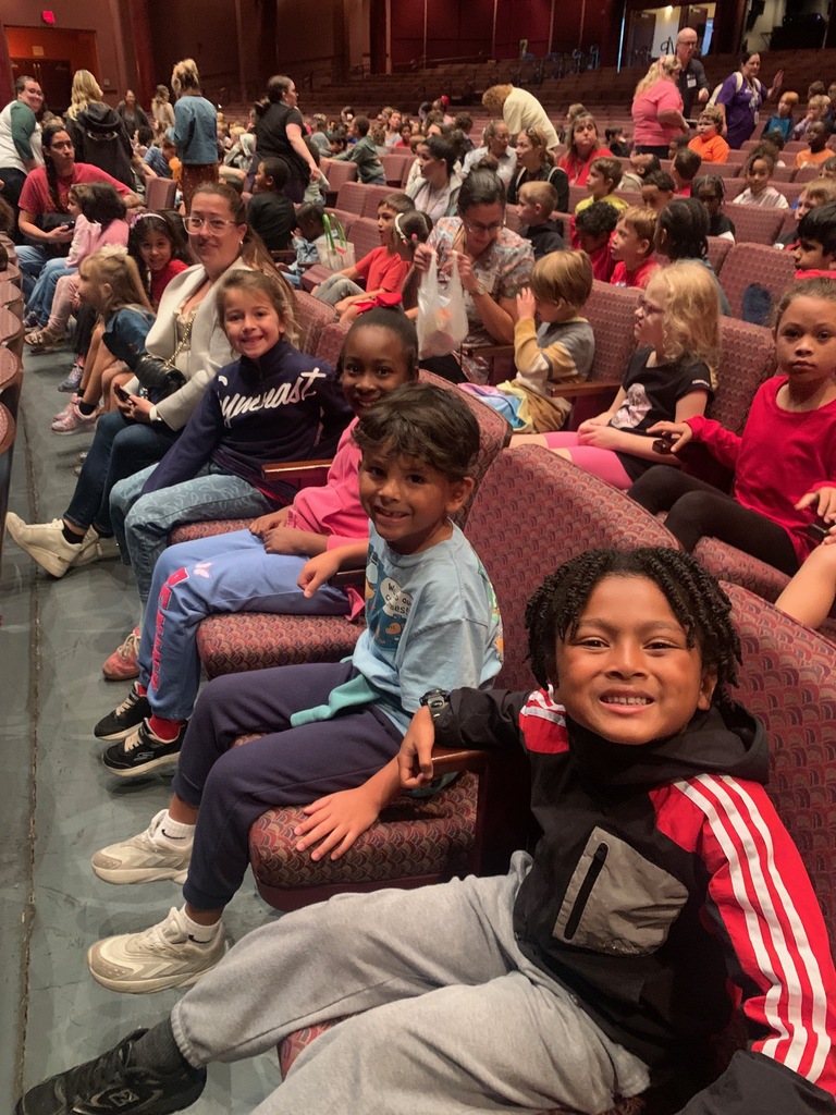 Group of children seated in an auditorium. Some hold hands, others wear backpacks. Row of chairs and dark wall behind them.