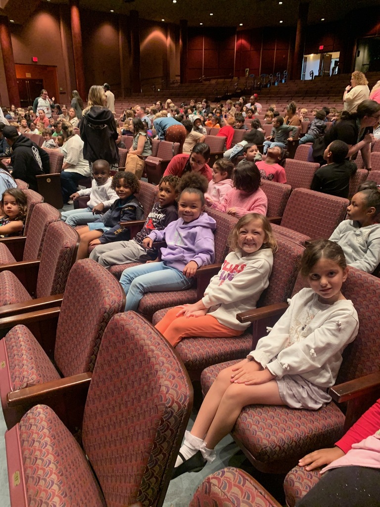 Many children in various clothing sit in rows of red chairs, facing a stage. Some adults sit in the back.