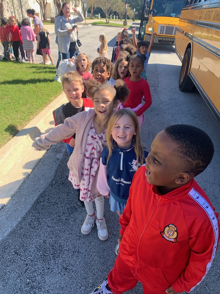 Group of young children standing outside by a school bus on a road, with others in background