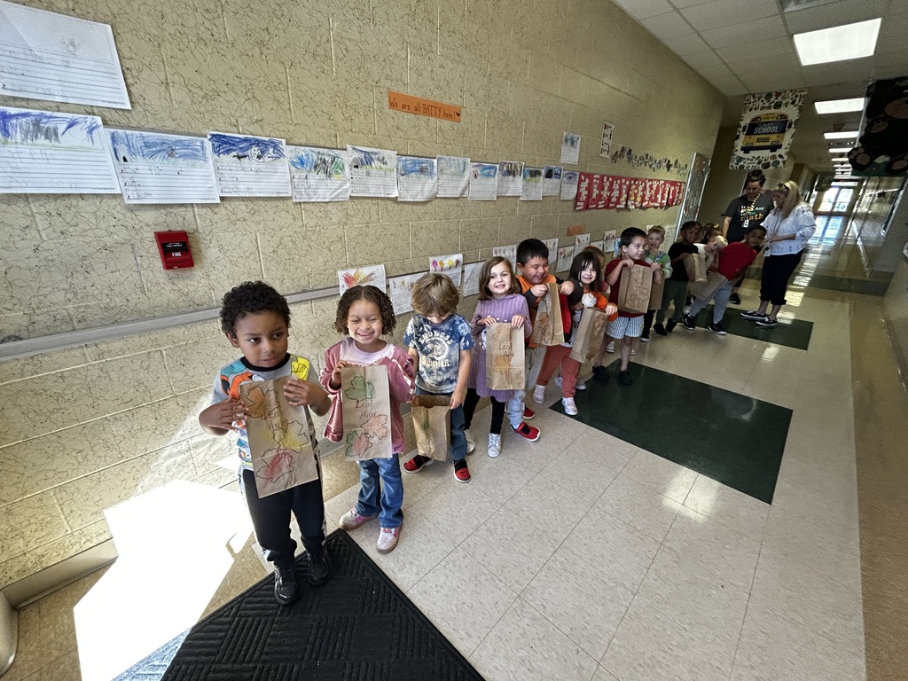 Children holding paper bags lined up in a hallway. Papers are attached to the wall.