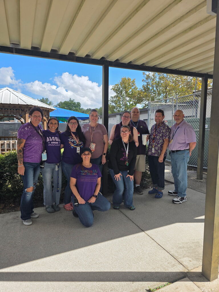 Nine people pose for a photo under an awning, with some crouching and others standing, in a shaded area.
