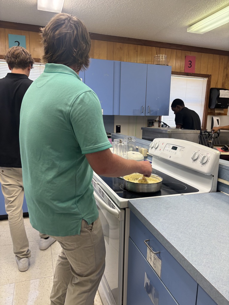 Two students prepare food in a kitchen; one stirs a pan on a stove, another watches.