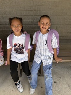 Two children standing next to each other wearing backpacks, smiling and looking at the camera.