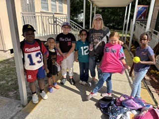 Several children stand in line outside a school building. A girl holds a ball. A woman stands with them.