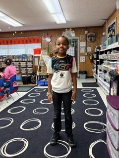 A child stands on a black and white rug in a classroom. Behind them are shelves, a fire extinguisher, and a person sitting in a chair.