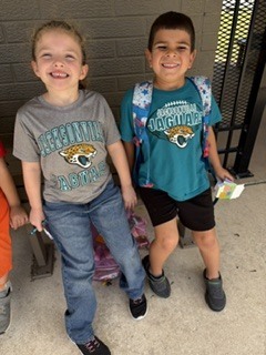 Two boys standing next to each other. One wears a gray t-shirt with a tiger design. The other wears a teal shirt and carries a backpack.