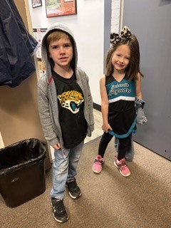 Two children stand in a hallway; one wears a hoodie and the other a jersey with a bow.