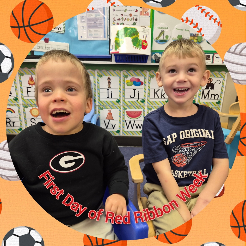 Two young boys sit together, smiling. A red ribbon is around one boy's neck. They sit on chairs with a colorful background of sports balls.