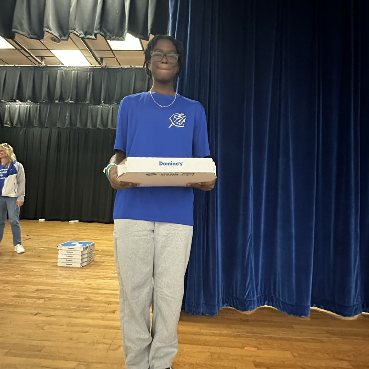 Person standing on stage holding box, blue curtain behind, another person in background, and boxes on floor.