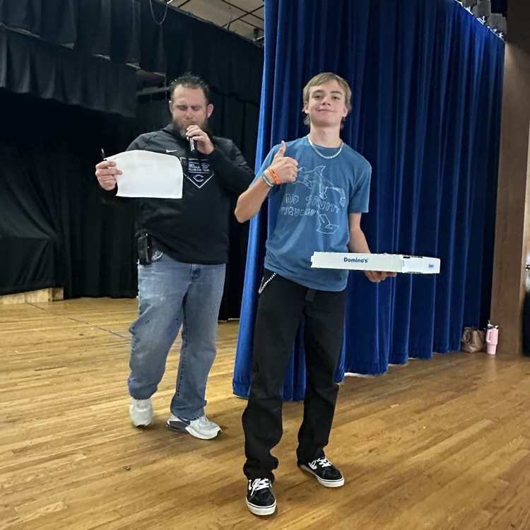 Two people stand on a stage; one holds a pizza box, the other a paper and microphone.