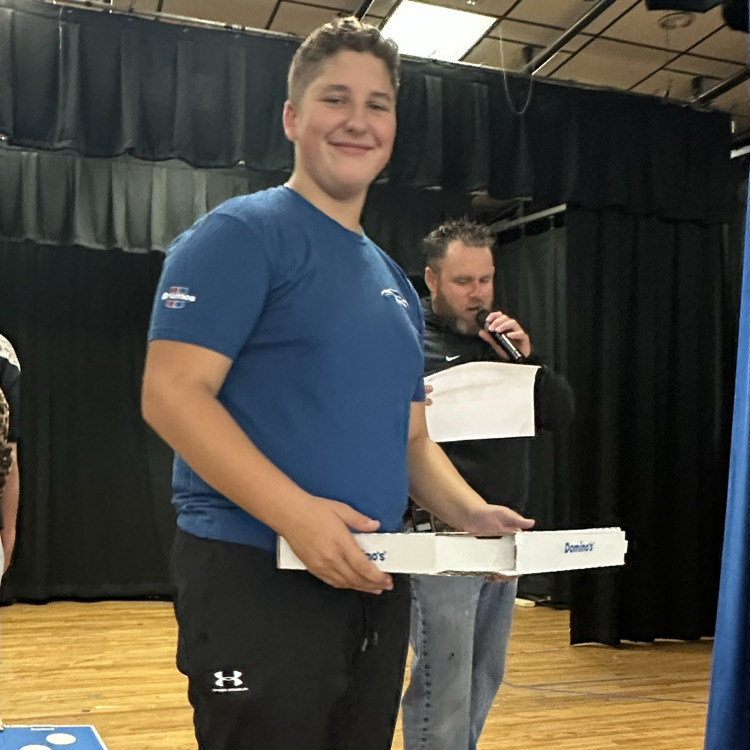 Two people hold boxes of pizza; one holds a microphone. They stand on a stage with black curtains behind them.