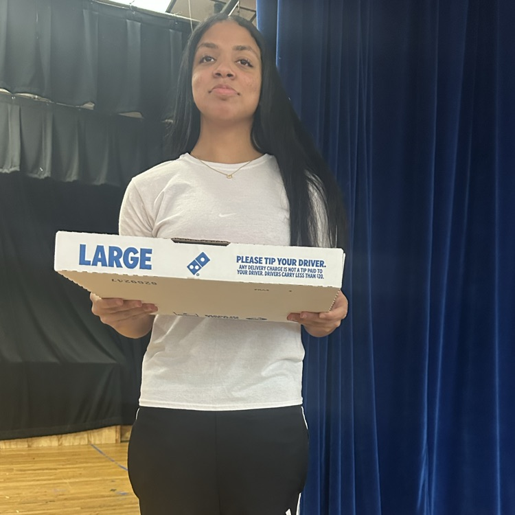 Woman wearing white T-shirt and black pants holding a large pizza box in front of a blue curtain.