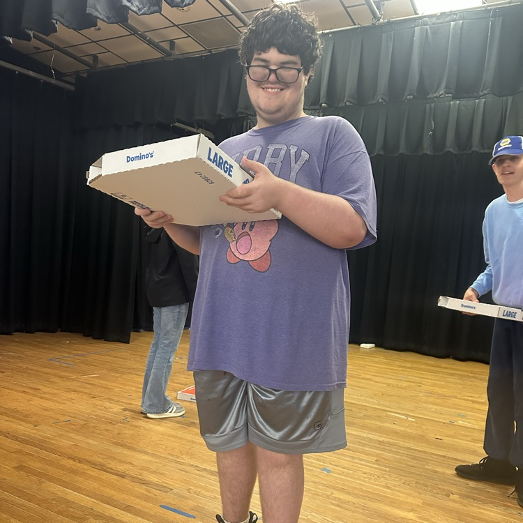 A student holds a large pizza box while standing on a stage in a room with a black backdrop.