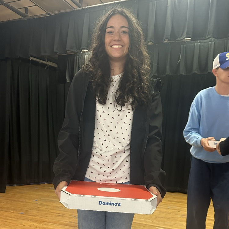 Person holds pizza box with a smiling face; another holds paper and stands behind them on a stage.