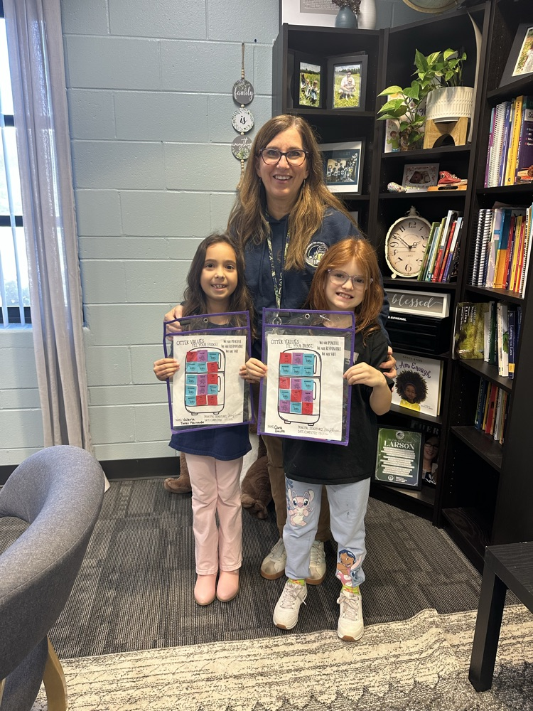 Three children hold awards in a room with bookshelves. A woman wearing glasses stands between two young girls.