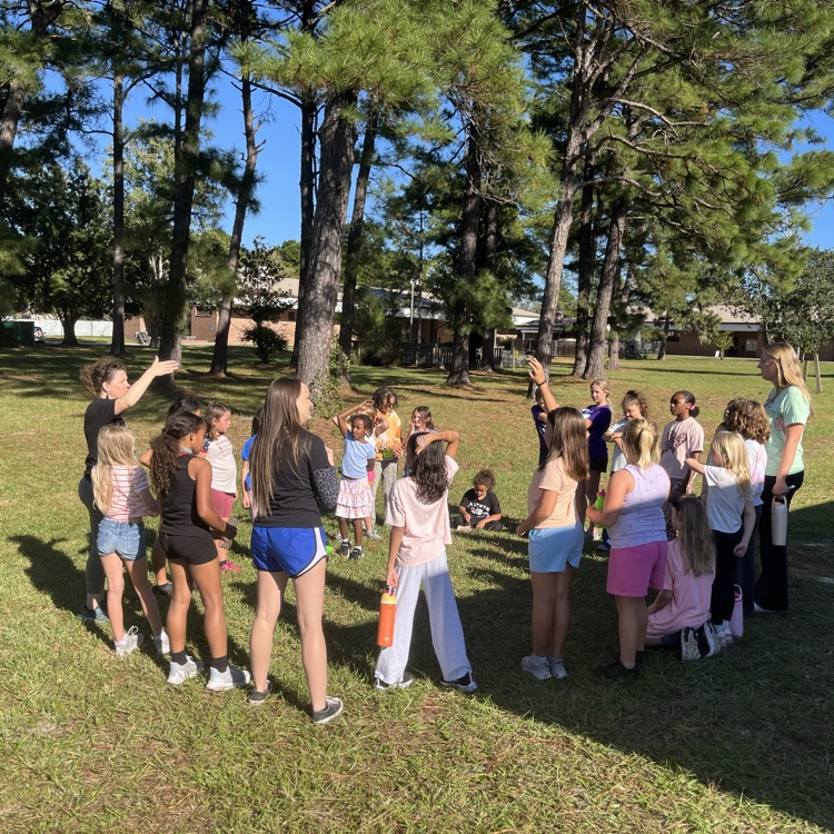 Group of students outside with adults; some are standing, some are crouched; one adult points at something