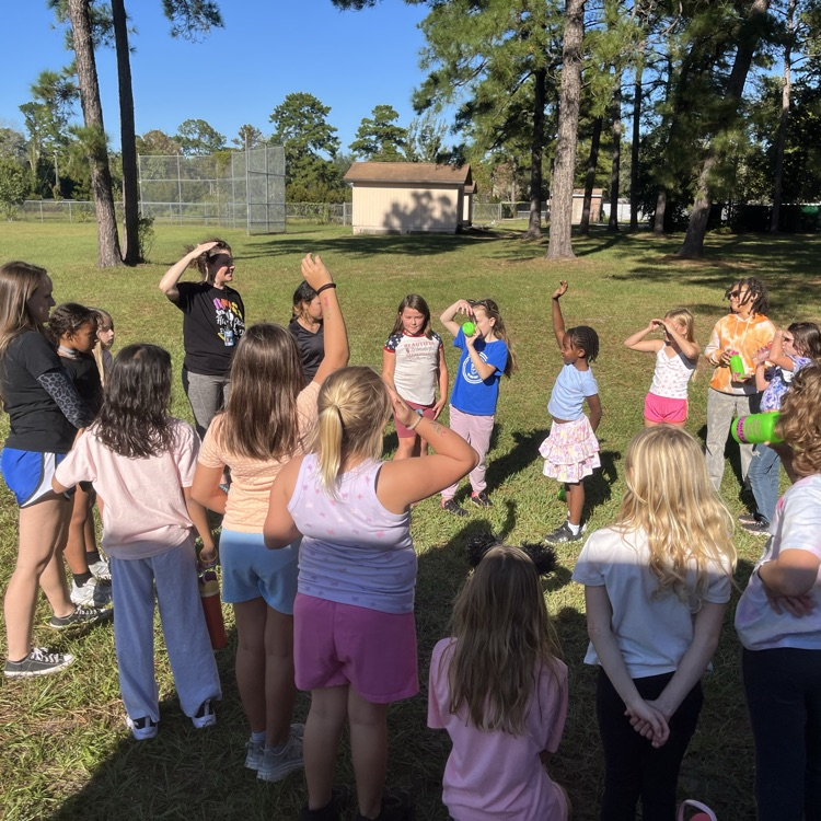 Several children and adults are gathered in a grassy field, some raising their hands.