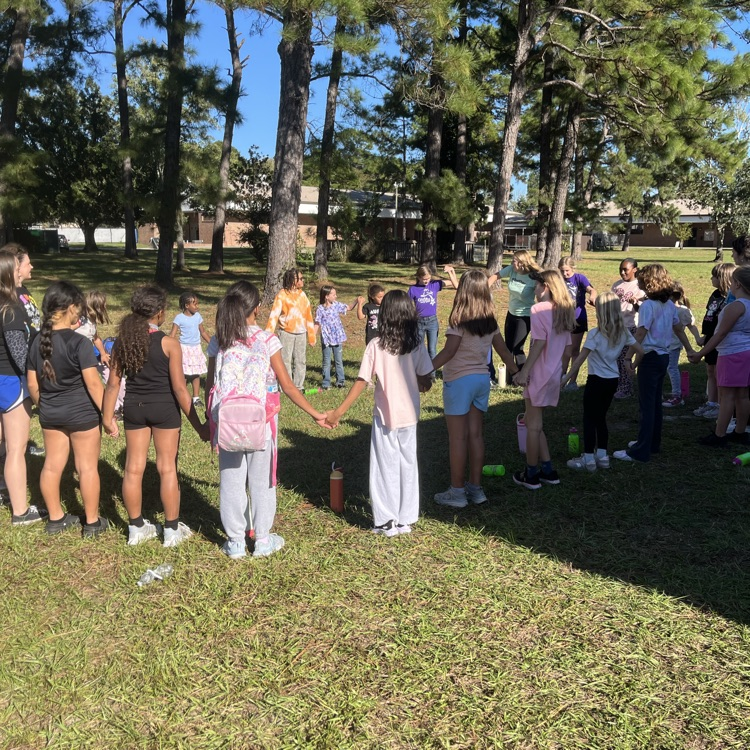 Students in shorts and t-shirts standing in a circle with hands held in the center.