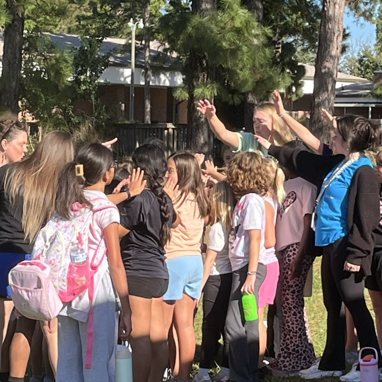 A group of children and adults stand in a circle in a grassy area, some with backpacks.