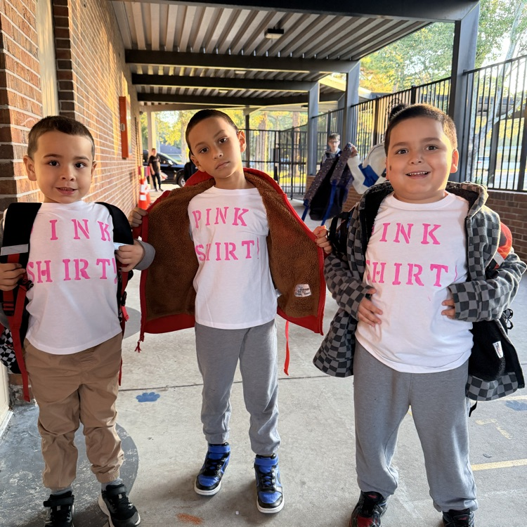 Three boys wearing pink shirts and backpacks standing in a school corridor. The middle boy holds a red jacket.