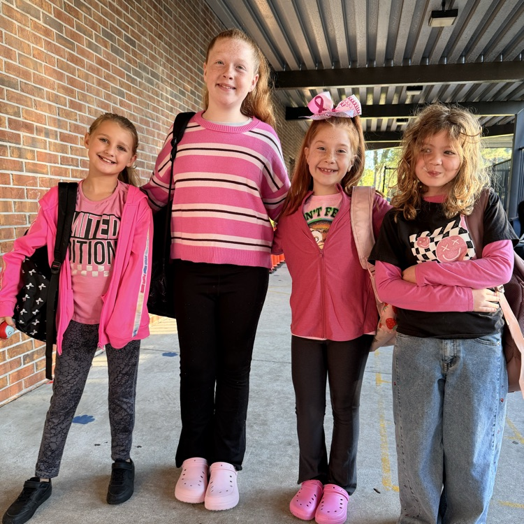 Four girls standing together wearing pink shoes, pink jackets, and carrying backpacks. A brick wall in the background.