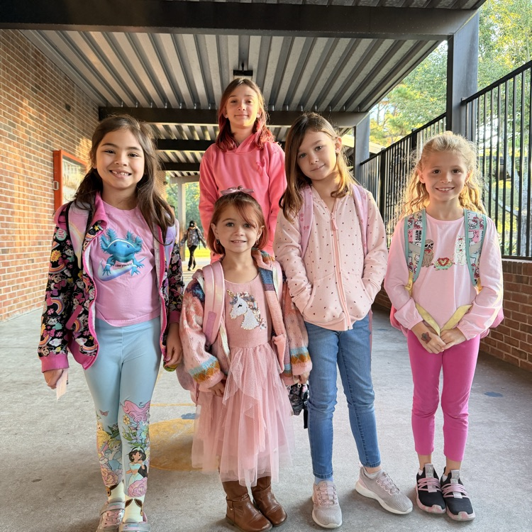 Five girls in matching pink tops, backpacks, and colorful pants pose in a hallway.