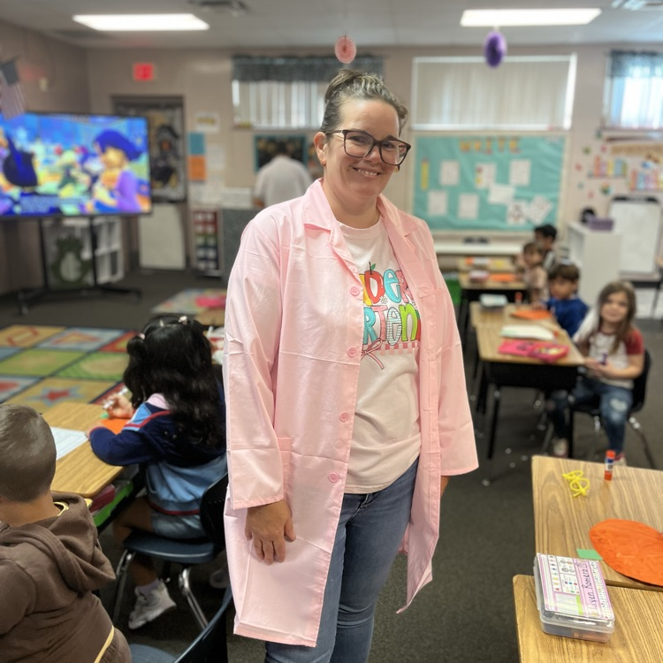 A teacher stands in a classroom with students seated at desks. She wears a pink coat and glasses.