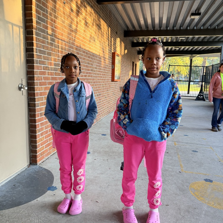 Two girls stand in a hallway. They wear jackets, gloves, and pink pants. One has a pink backpack.