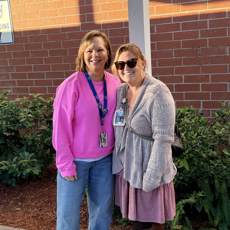 Two women stand on a concrete sidewalk in front of a brick building. One wears a pink sweater and jeans, the other a gray sweater and brown boots.