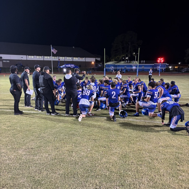 Blue-uniformed players kneel on a grass field. Coaches and officials stand nearby. A scoreboard shows the time.