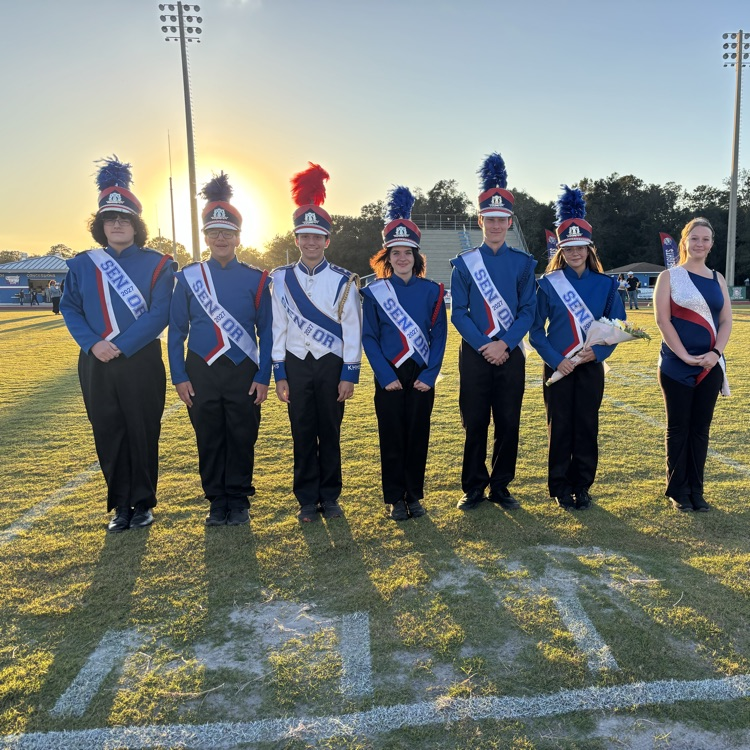 Students in uniforms, with hats and flowers, stand on a field, with white lettering on the grass.