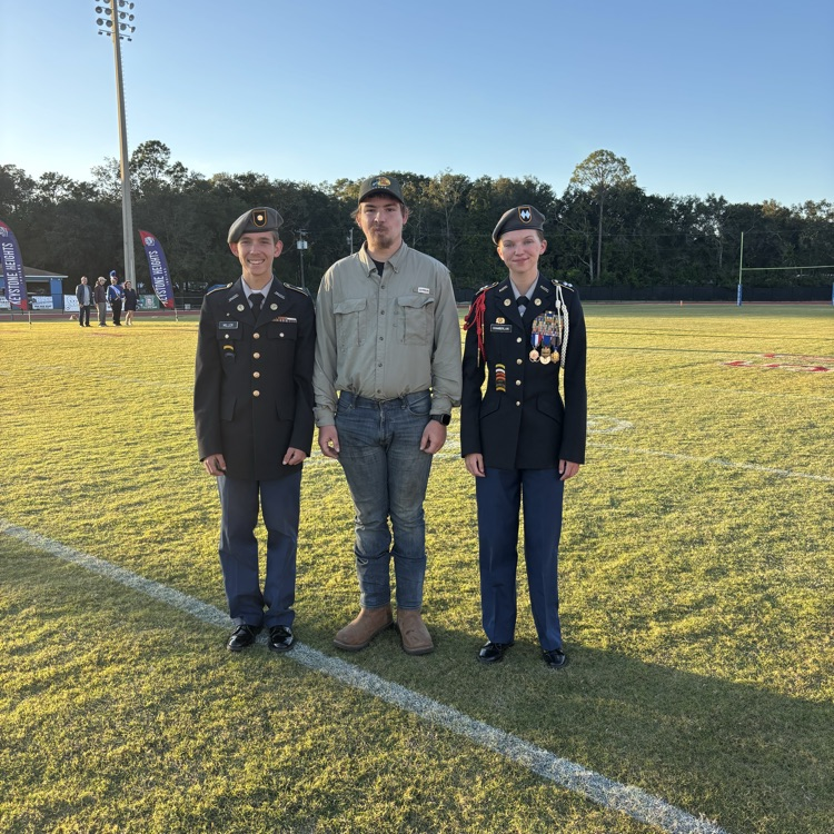 Three people stand on a grassy field with white lines. Two in uniforms, one in casual attire. Trees and a pole in the distance.