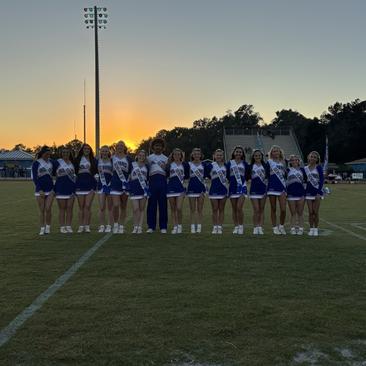 Cheerleaders in blue and white uniforms stand on a field with white lines, near a streetlight.