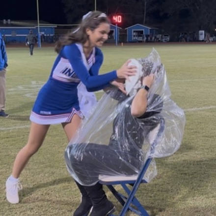 Two people on a field. A girl in a blue uniform helps a seated person. Another person stands behind.