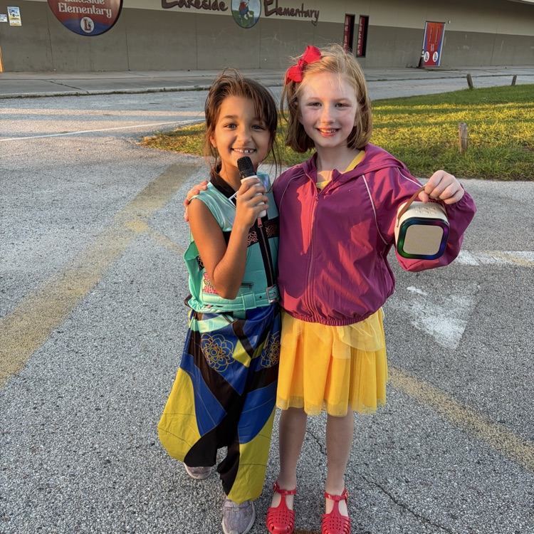 Two girls standing in a parking lot. One holds a microphone, and the other holds a bag.