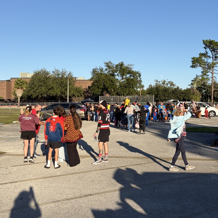 Many people stand in an outdoor parking lot with trees and buildings in the background.