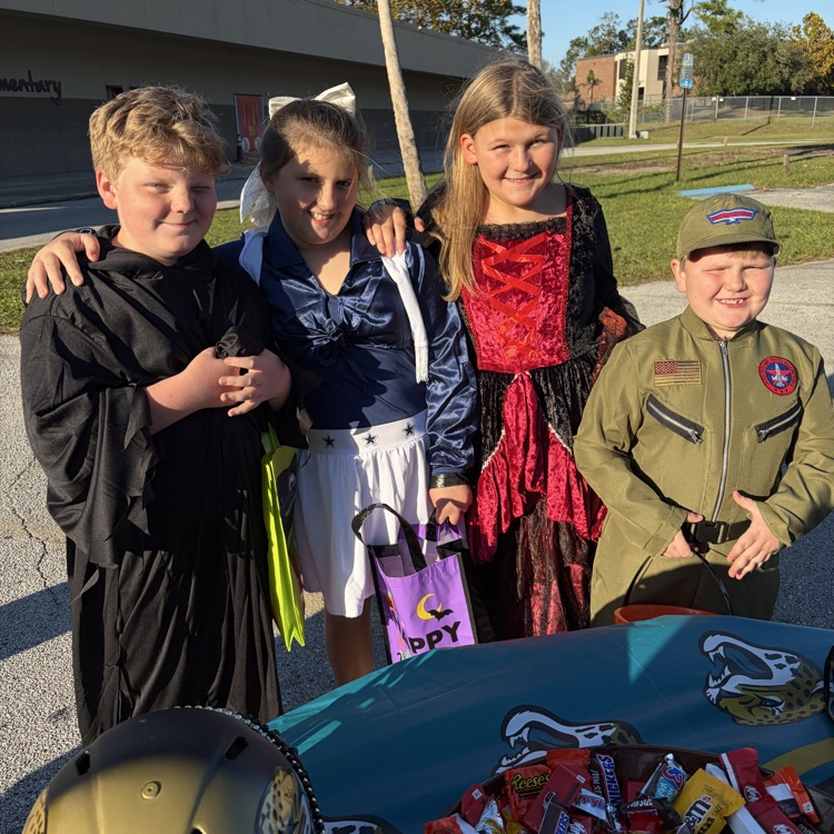 Four children in costumes, two boys and two girls, standing together. One has a red cape, another a blue jacket. Two tables with candy and a helmet.