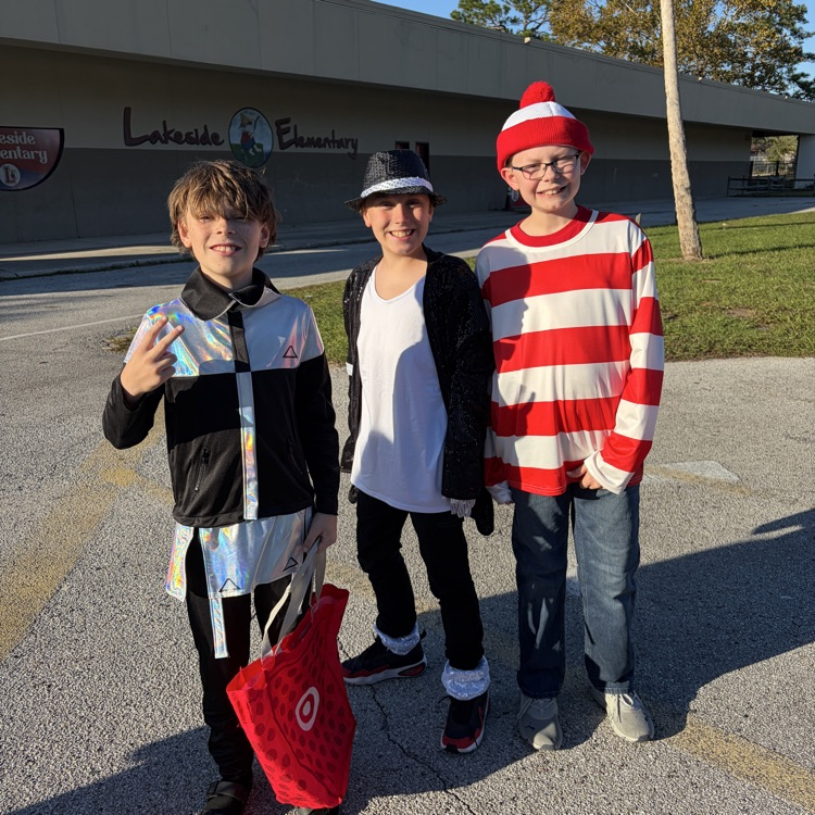 Three boys in Halloween costumes stand in a parking lot. The boy in the middle holds a red bag.