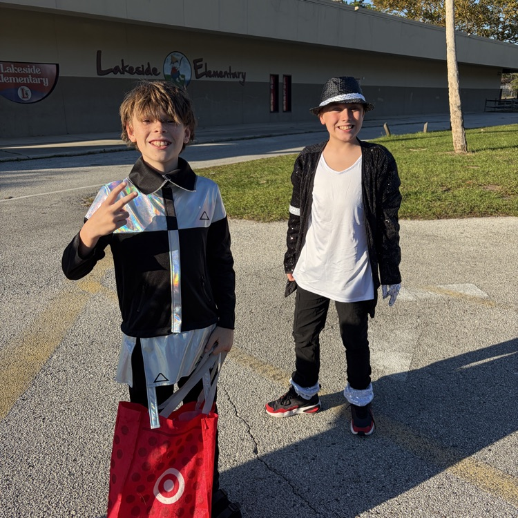 Two young boys in matching outfits stand by the curb outside a school building.