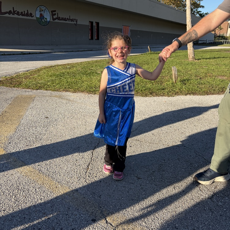 Young girl in cheerleading uniform and glasses holds hand of adult wearing green pants and black shoes, standing outside near school building.