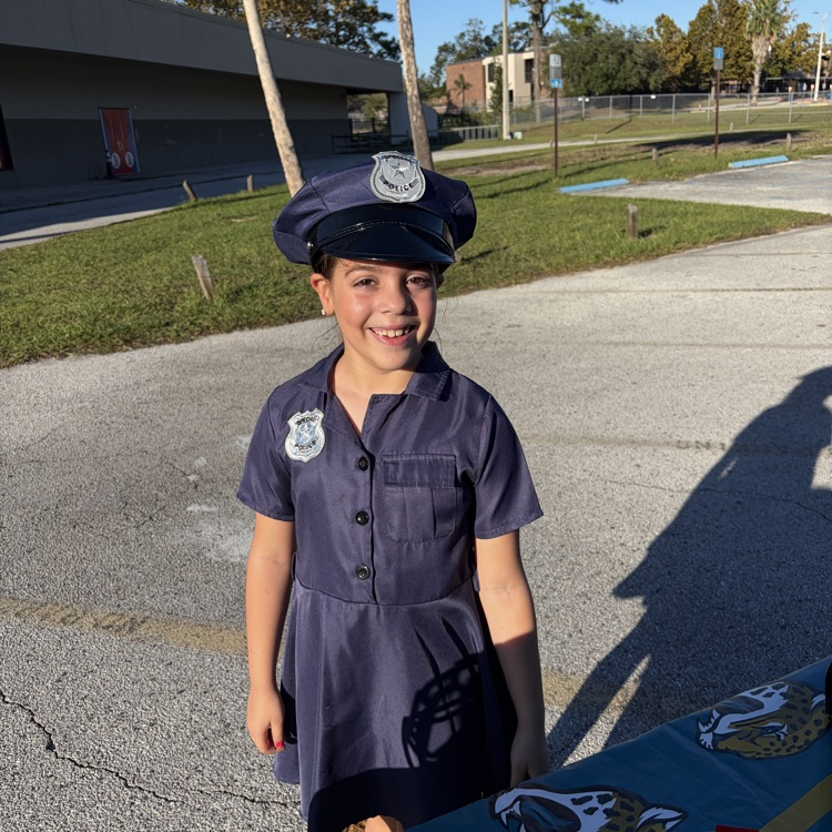 Young child in police uniform stands near a table with candy, smiling, in a parking lot.